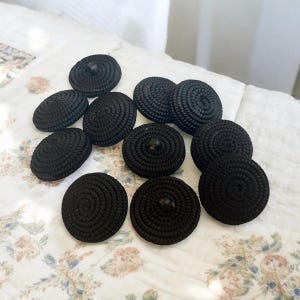 Pu&ograve; includere: Una collezione di 11 bottoni neri con un design a spirale. I bottoni sono realizzati in materiale tessuto e hanno un gambo in plastica nera.