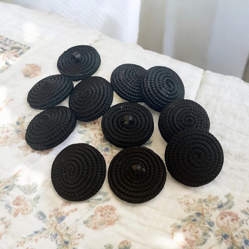 Pu&ograve; includere: Una collezione di 11 bottoni neri con un design a spirale. I bottoni sono realizzati in materiale tessuto e hanno un gambo in plastica nera.