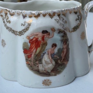 May include: White porcelain creamer with a gold trim and a painted scene of three women in a garden. The creamer has a fluted design and a handle.