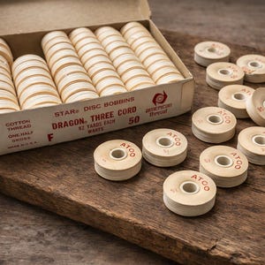 May include: A box of vintage thread spools labeled "STAR DISC BOBBINS DRAGON THREE CORD." The box contains multiple spools, with additional loose spools scattered on a wooden surface. The spools are cream-colored with a central hole.