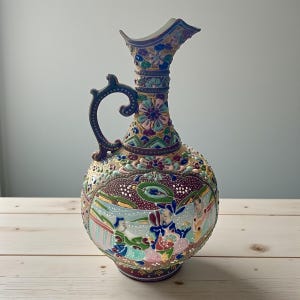 May include: A decorative ceramic pitcher with a handle, featuring a detailed floral and figural design. The pitcher has a bulbous body and a tall, fluted neck. The color palette includes shades of blue, green, red, and gold.