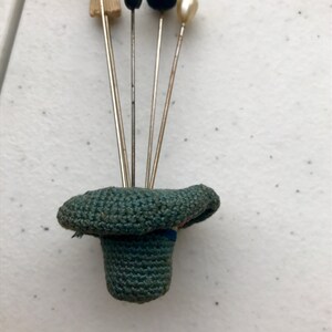 May include: A collection of hat pins with various decorative tops, including a blue fabric bow, a tan brush, a black bead, a black pom-pom, and a pearl. The pins are displayed in a small, green crocheted hat.