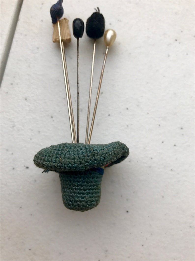 May include: A collection of hat pins with various decorative tops, including a blue fabric bow, a tan brush, a black bead, a black pom-pom, and a pearl. The pins are displayed in a small, green crocheted hat.