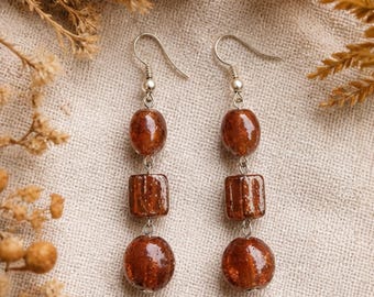 May include: A pair of dangle earrings with a silver hook and three amber-colored beads. The beads are a mix of oval, square, and round shapes. The earrings are displayed on a beige fabric background.