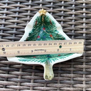 May include: A ceramic Christmas tree-shaped dish, approximately 6 inches long, with hand-painted green branches, red and blue ornaments, and a gold star on top. The dish is on a woven surface.