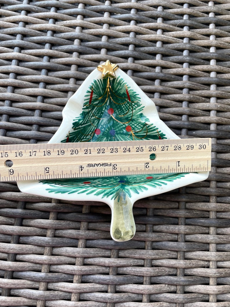 May include: A ceramic Christmas tree-shaped dish, approximately 6 inches long, with hand-painted green branches, red and blue ornaments, and a gold star on top. The dish is on a woven surface.
