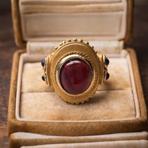 May include: A gold-toned ring with a large, oval, deep red gemstone at its center. The ring has intricate detailing, including a rope-like border around the gemstone and small blue stones on each side. The ring is displayed in a velvet-lined jewelry box.