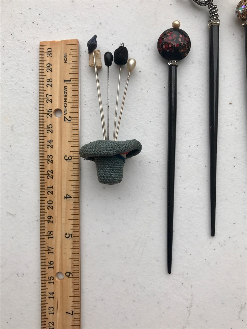 May include: A collection of vintage hat pins and hair sticks. The hat pins feature various decorative tops, including a small bird, black beads, and a pearl. A ruler shows the pins are approximately 3 inches (7.6 cm) tall.