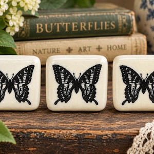 May include: Three square, off-white ceramic knobs, each featuring a detailed black butterfly silhouette printed on the front surface. The knobs have rounded edges and a smooth, glossy finish, displaying a consistent design across all three pieces.