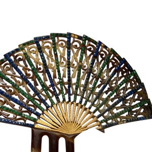 May include: A decorative hair comb in a fan shape. The comb features an openwork design with gold-colored metal and is adorned with blue and green gemstones. The base of the comb is a dark brown color.