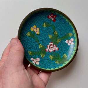 May include: A small, round, blue and green enamel trinket dish with a floral design. The dish has a brass rim and is decorated with pink, yellow, and red flowers.