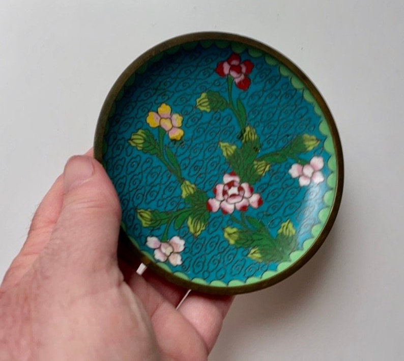 May include: A small, round, blue and green enamel trinket dish with a floral design. The dish has a brass rim and is decorated with pink, yellow, and red flowers.