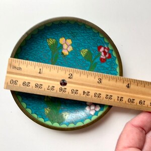 May include: A small, round, blue and green enamel trinket dish with a floral design. The dish has a gold rim and is approximately 7.5 cm in diameter.
