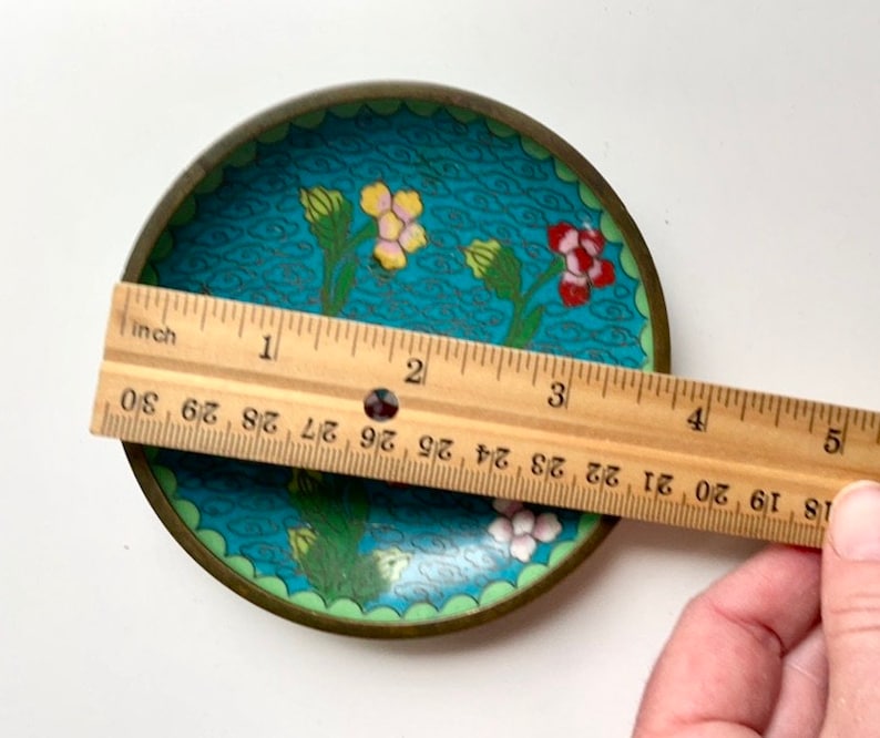 May include: A small, round, blue and green enamel trinket dish with a floral design. The dish has a gold rim and is approximately 7.5 cm in diameter.