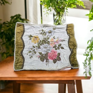 May include: A decorative pillow with a floral design featuring pink, white, and yellow roses. The pillow has a light-colored background with a green and brown border. It is placed on a wooden table.