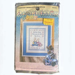 May include: A sealed cross-stitch sampler kit from Daisy Kingdom, featuring a baby-themed design. The sampler includes a framed cross-stitch with two bunnies, the name "Jacklyn Anne", and the birthdate of February 18, 1991, and weight of 7 lbs. 10 oz.