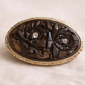 May include: Oval-shaped, dark brown carved brooch with a floral design and two clear crystal accents. The brooch is framed by a gold-colored, ornate metal border. The carved details and crystal accents add a touch of elegance.