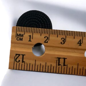 Pu&ograve; includere: Un bottone nero con una superficie testurizzata, misurato con un righello. Il bottone ha un diametro di circa 2,5 cm.