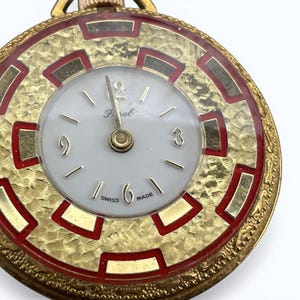 May include: A gold-toned pocket watch with a white face and gold hands. The watch has a textured gold bezel with red accents and a gold chain. The watch face has the word "Perol" and "SWISS MADE" printed on it.