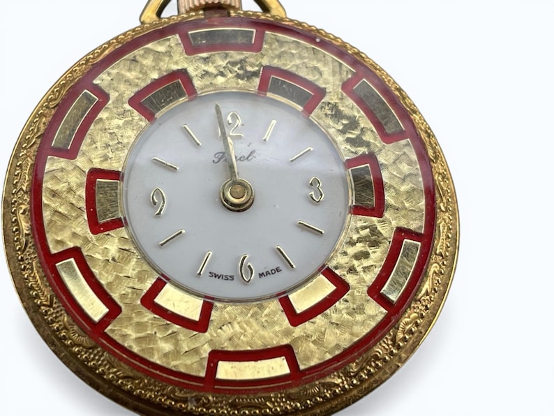 May include: A gold-toned pocket watch with a white face and gold hands. The watch has a textured gold bezel with red accents and a gold chain. The watch face has the word "Perol" and "SWISS MADE" printed on it.