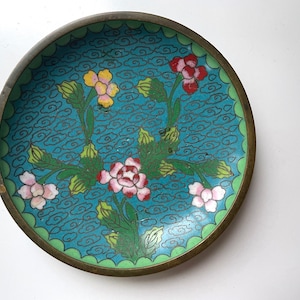 May include: A small, round, brass plate with a blue enamel background and a floral design. The flowers are pink, white, yellow, and red. The plate has a green border.
