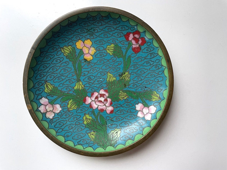 May include: A small, round, brass plate with a blue enamel background and a floral design. The flowers are pink, white, yellow, and red. The plate has a green border.