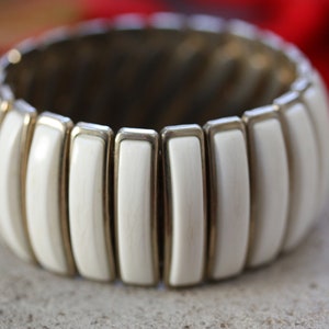 Peut inclure: Bracelet extensible ivoire avec un design côtelé. Le bracelet présente des segments rectangulaires avec des accents métalliques dorés. Le bijou est posé sur un fond neutre avec un élément rouge flou.