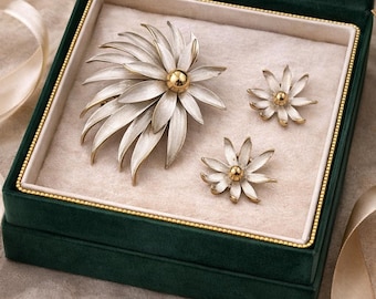 May include: A vintage jewellery set comprising a large flower brooch and two matching earrings, displayed in a green velvet box. The brooch and earrings are white and gold, with a detailed floral design.