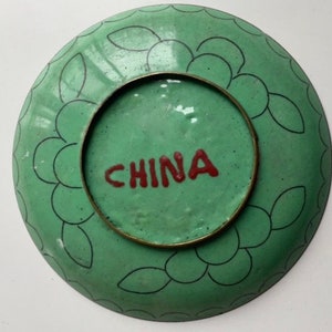 May include: A green ceramic plate with a black and white floral design around the edge. The plate has a brass rim and the word "CHINA" is written in red on the bottom.
