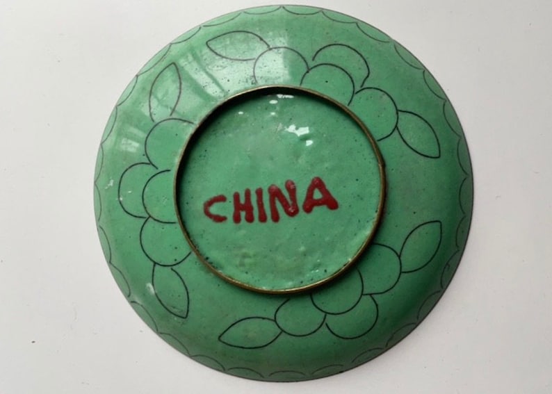 May include: A green ceramic plate with a black and white floral design around the edge. The plate has a brass rim and the word "CHINA" is written in red on the bottom.