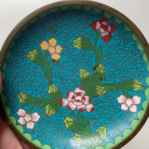 May include: A small, round, blue and green enamel dish with a floral design. The dish has a brass rim and is decorated with stylised flowers and clouds.