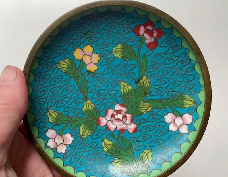 May include: A small, round, blue and green enamel dish with a floral design. The dish has a brass rim and is decorated with stylised flowers and clouds.