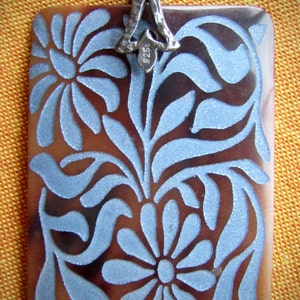 May include: A silver pendant with a rectangular brown and blue design. The pendant features a floral pattern with a silver bail.