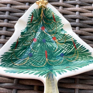 May include: A ceramic Christmas tree-shaped dish with a gold star on top. The tree is painted green with red and blue ornaments and gold garland. The dish has a white base and a handle.