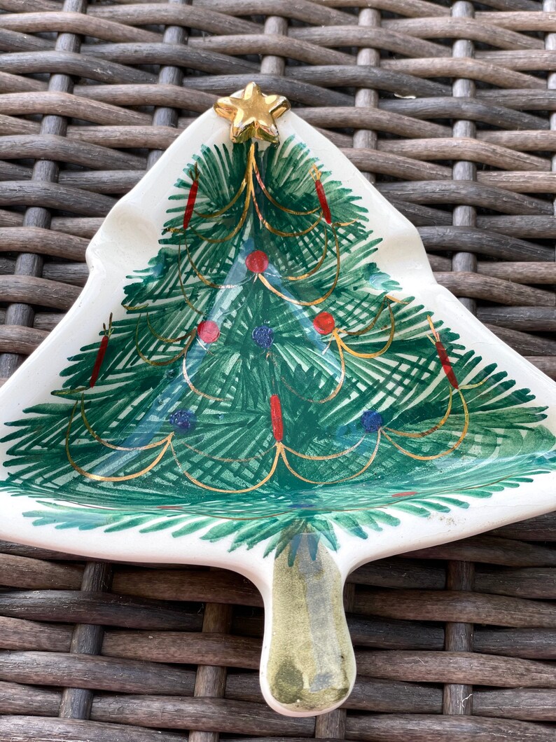 May include: A ceramic Christmas tree-shaped dish with a gold star on top. The tree is painted green with red and blue ornaments and gold garland. The dish has a white base and a handle.