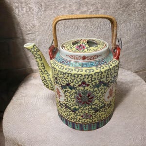 May include: A yellow porcelain teapot with a bamboo handle and intricate floral and geometric designs. The teapot has a lid with two holes and features Chinese characters.
