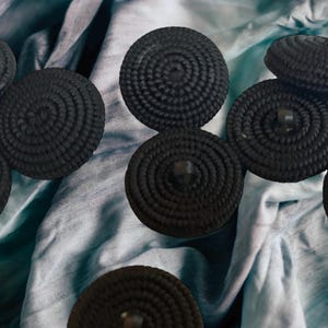 Pu&ograve; includere: Una collezione di otto bottoni rotondi neri intrecciati. I bottoni sono disposti su uno sfondo di tessuto blu e bianco.