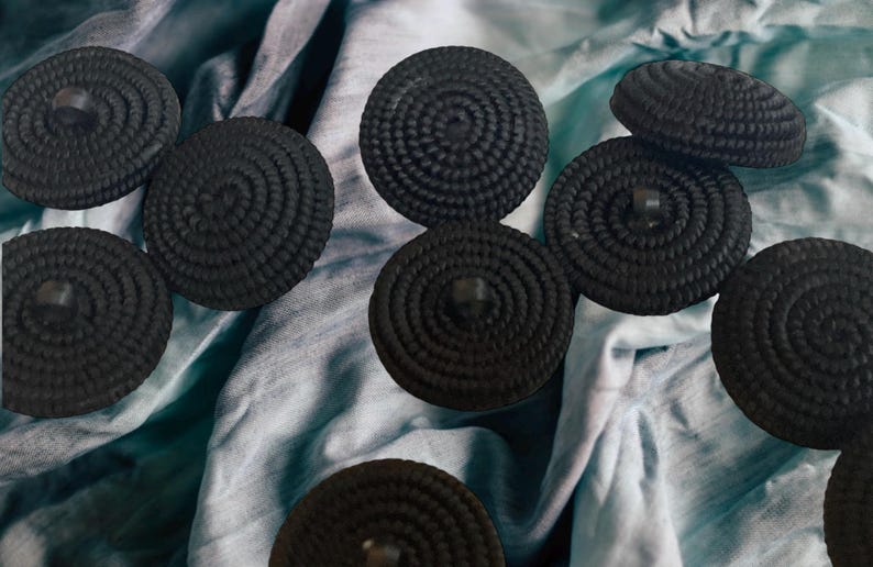 Pu&ograve; includere: Una collezione di otto bottoni rotondi neri intrecciati. I bottoni sono disposti su uno sfondo di tessuto blu e bianco.
