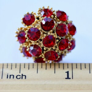 May include: A close-up of a red rhinestone ball ornament measuring 1 inch in diameter. The ball is made of many small red rhinestones set in gold metal.