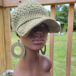 May include: A crocheted olive green hat with a visor and matching large hoop earrings. The hat and earrings are displayed on a mannequin head. The earrings are circular and have a thick, textured appearance. The hat has a textured pattern.