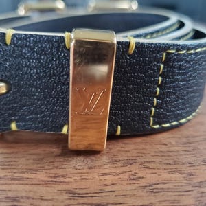 May include: A black leather belt with yellow stitching and a gold buckle featuring the Louis Vuitton logo.