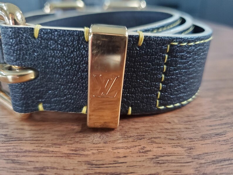 May include: A black leather belt with yellow stitching and a gold buckle featuring the Louis Vuitton logo.