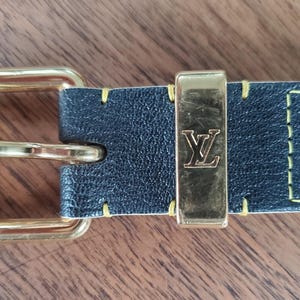 May include: A black leather belt with a gold buckle and a yellow stitching detail. The buckle has the letter "L" embossed on it.