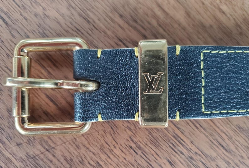 May include: A black leather belt with a gold buckle and a yellow stitching detail. The buckle has the letter "L" embossed on it.