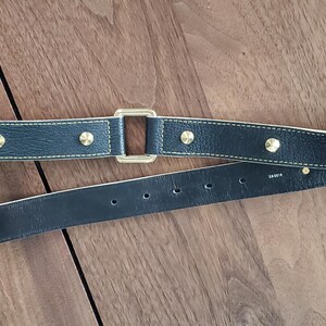 May include: A black leather belt with gold-toned hardware and studs. The belt has a buckle on one end and a square-shaped piece of hardware on the other end.