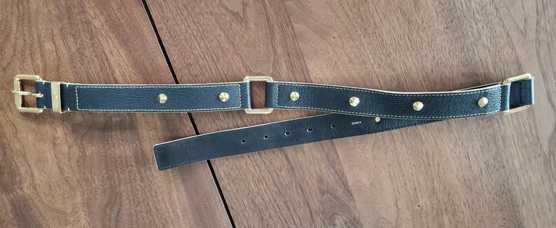 May include: A black leather belt with gold-toned hardware and studs. The belt has a buckle on one end and a square-shaped piece of hardware on the other end.