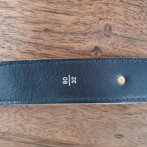 May include: Close-up of a black leather belt with white stitching and a gold metal buckle. The belt is labeled "80/32".