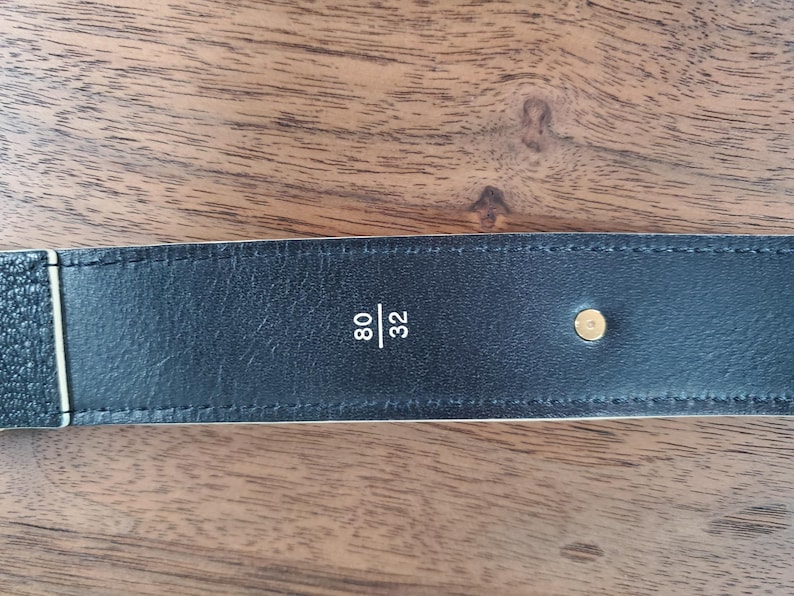 May include: Close-up of a black leather belt with white stitching and a gold metal buckle. The belt is labeled "80/32".