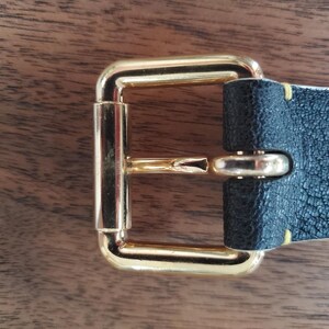 May include: A gold metal belt buckle with a black leather strap. The buckle has a rectangular shape with a smooth, polished finish. The strap is stitched with yellow thread.