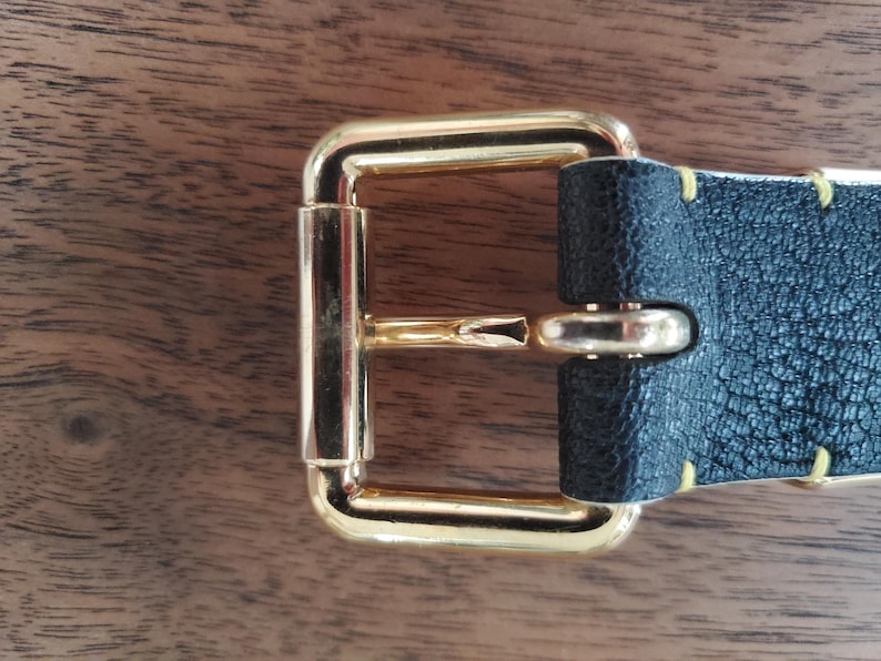 May include: A gold metal belt buckle with a black leather strap. The buckle has a rectangular shape with a smooth, polished finish. The strap is stitched with yellow thread.